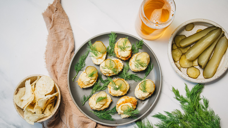 Overhead view of pickle and dill topped deviled eggs on a grey plate, surrounded by pickles, beer, and dill pickle chips.