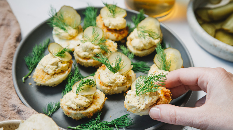 Hand lifting a pickle and dill topped deviled eggs from a grey plate.