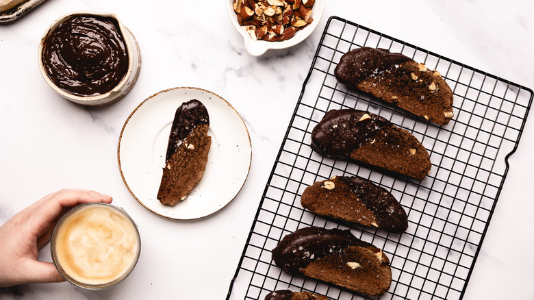 Four chocolate biscotti on a rack with hand holding coffee.