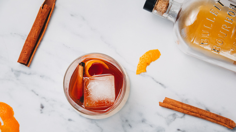 A glass of a brown butter old fashioned with bourbon bottle and cinnamon sticks