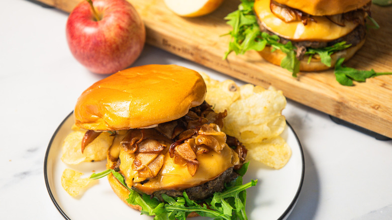 A caramelized apple burger on a plate with another one on a cutting board with apples